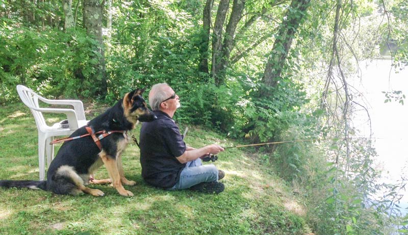 Man fishing on riverbank while his German shepherd guide dog watches over his shoulder
