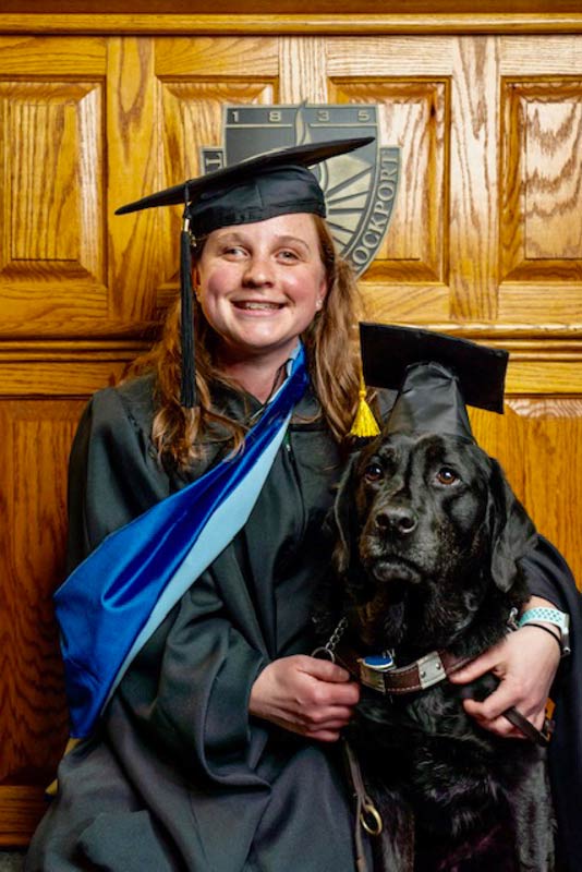 Woman in graduation gown hugging her black Lab guide dog, both are wearing caps.