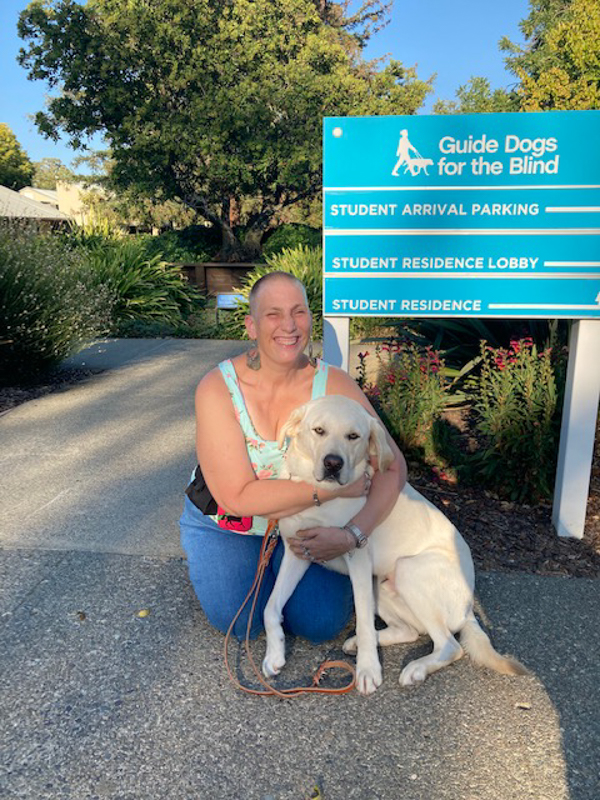 Woman hugging her yellow Lab in harness while kneeling next to a guide dog school sign