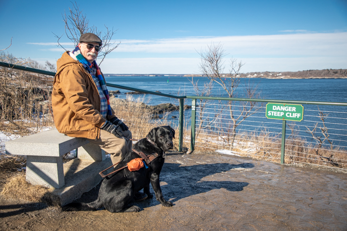 Brad sitting with his black Lab guide dog Stout at the ocean next to a sign that reads Danger, Steep Cliff