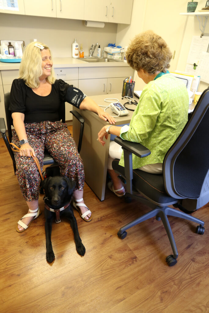 A nurse taking a blind woman’s blood pressure while her black Lab guide dog rests quietly under the chair.