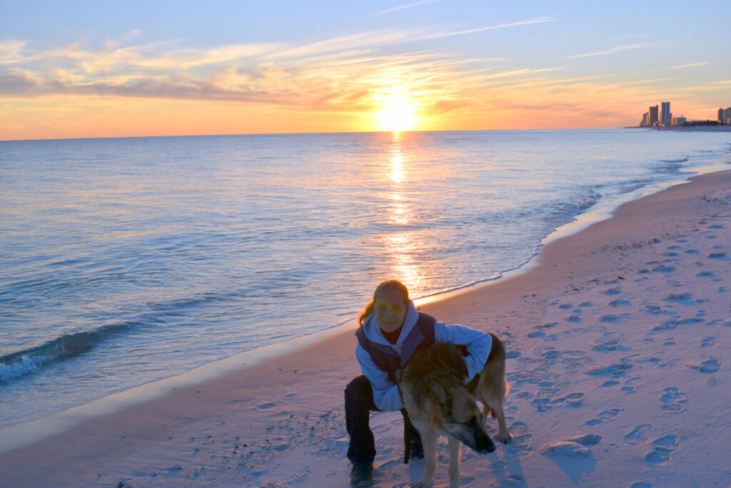 Sue is crouching on a beach at sunset with her German shepherd Seeing Eye dog beside her.