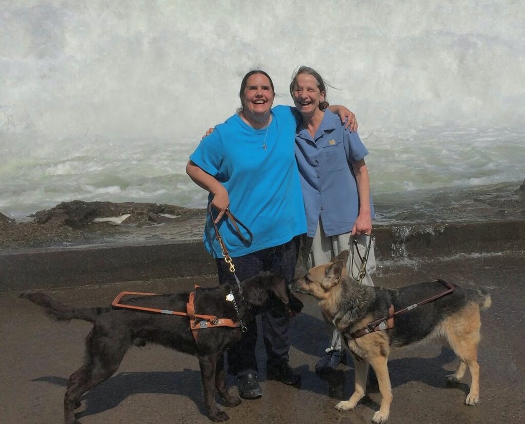 Two smiling friends with their arms around each other’s shoulders stand with their chocolate Lab and German Shepherd guide dogs in front of a large waterfall.