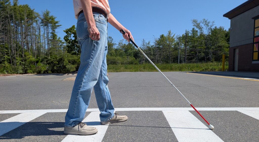 Close-up of a person using a long white cane while crossing a street in a marked crosswalk.