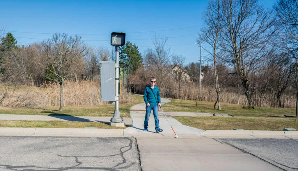 A young man with a white cane approaches a street crossing beneath a pedestrian signal.