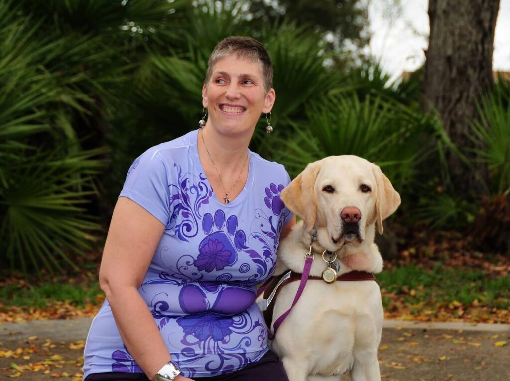 A smiling woman in a purple floral top sits beside her yellow Lab guide dog in a leafy outdoor setting.