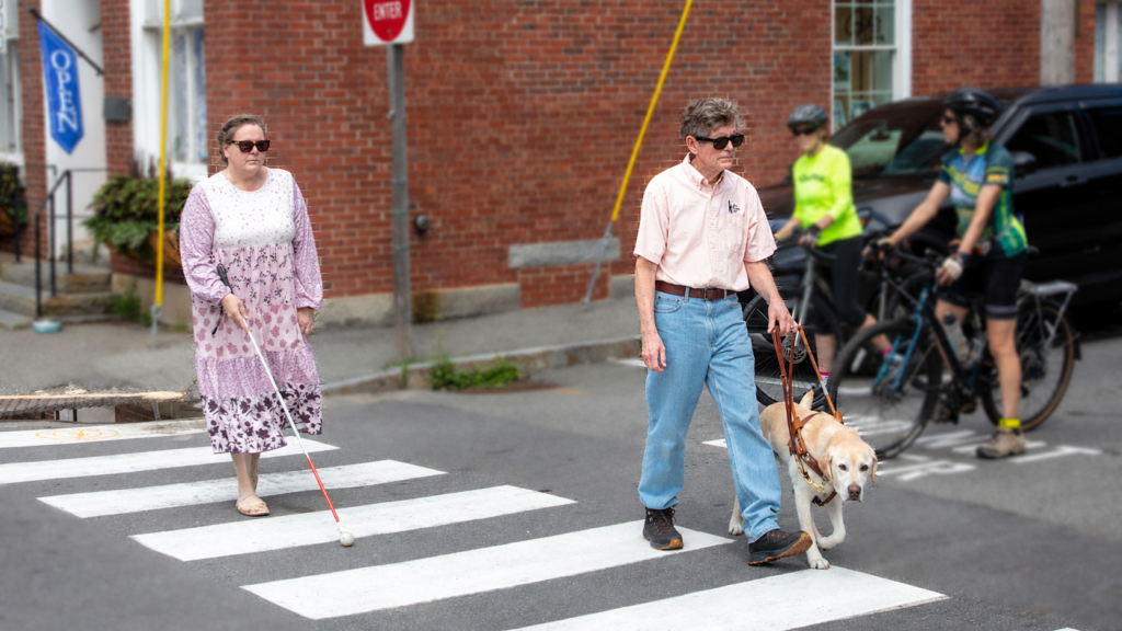 A woman using a white cane and a man with a yellow Lab guide dog crossing the street while two cyclists yield.