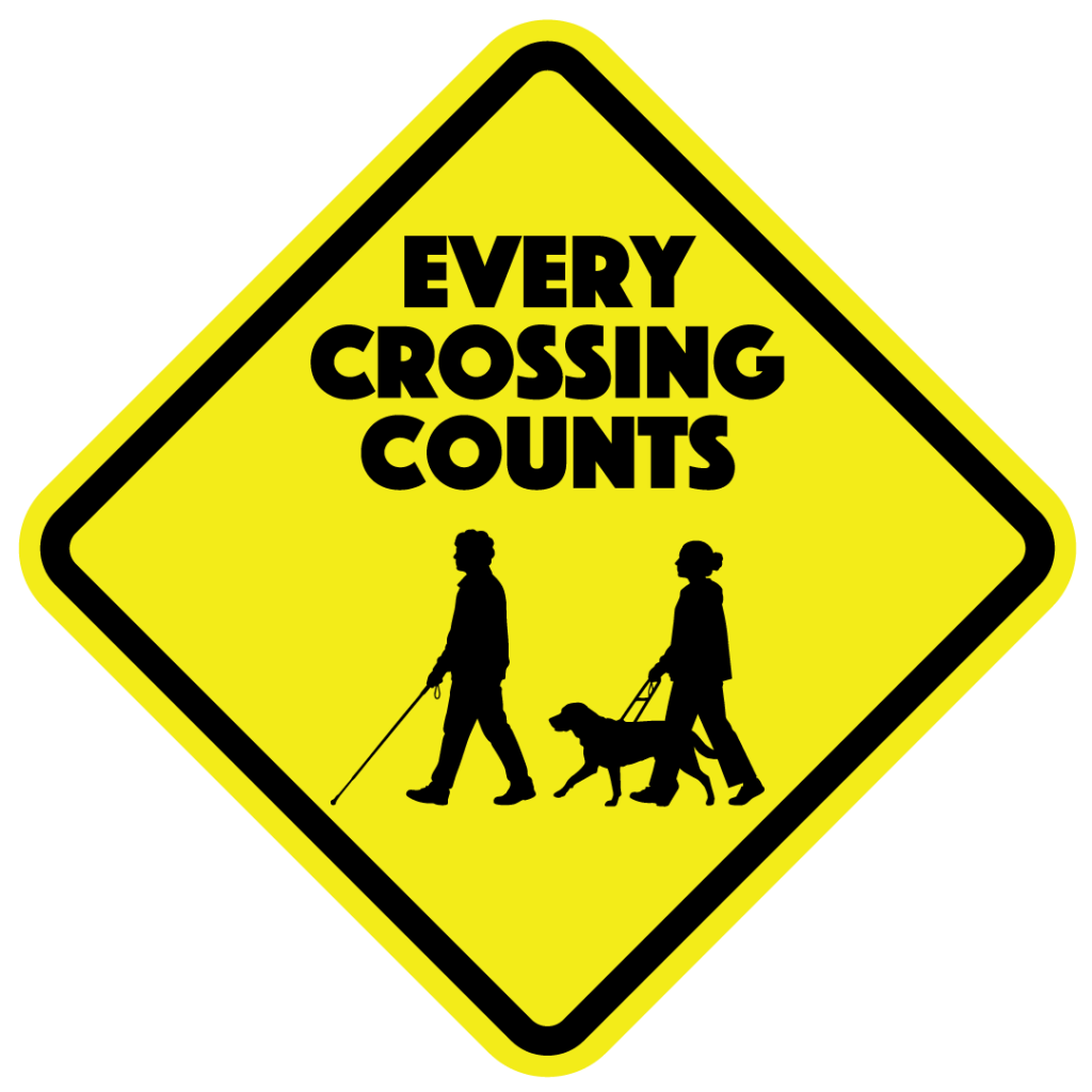 Every Crossing Counts campaign logo: a bright yellow diamond-shaped sign with a thick black border. Inside the sign, the words “Every Crossing Counts” are stacked above black silhouettes of a white cane user and a guide dog team walking in the same direction.