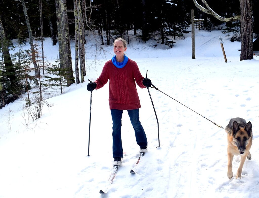 A woman in a red sweater and blue scarf cross-country skis while her off-duty German Shepherd guide dog trots alongside her.