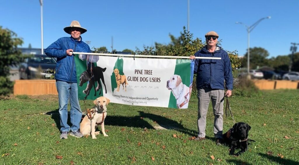 Two PTGDU members stand outdoors with a yellow Lab and a black Lab guide dog while holding the organization’s banner.