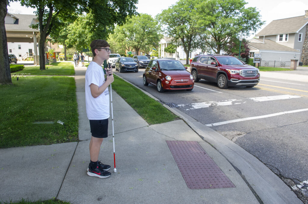 A young man wearing a bright blue polo shirt and shorts stands back from the edge of a marked crosswalk as several vehicles travel along the street in front of him.