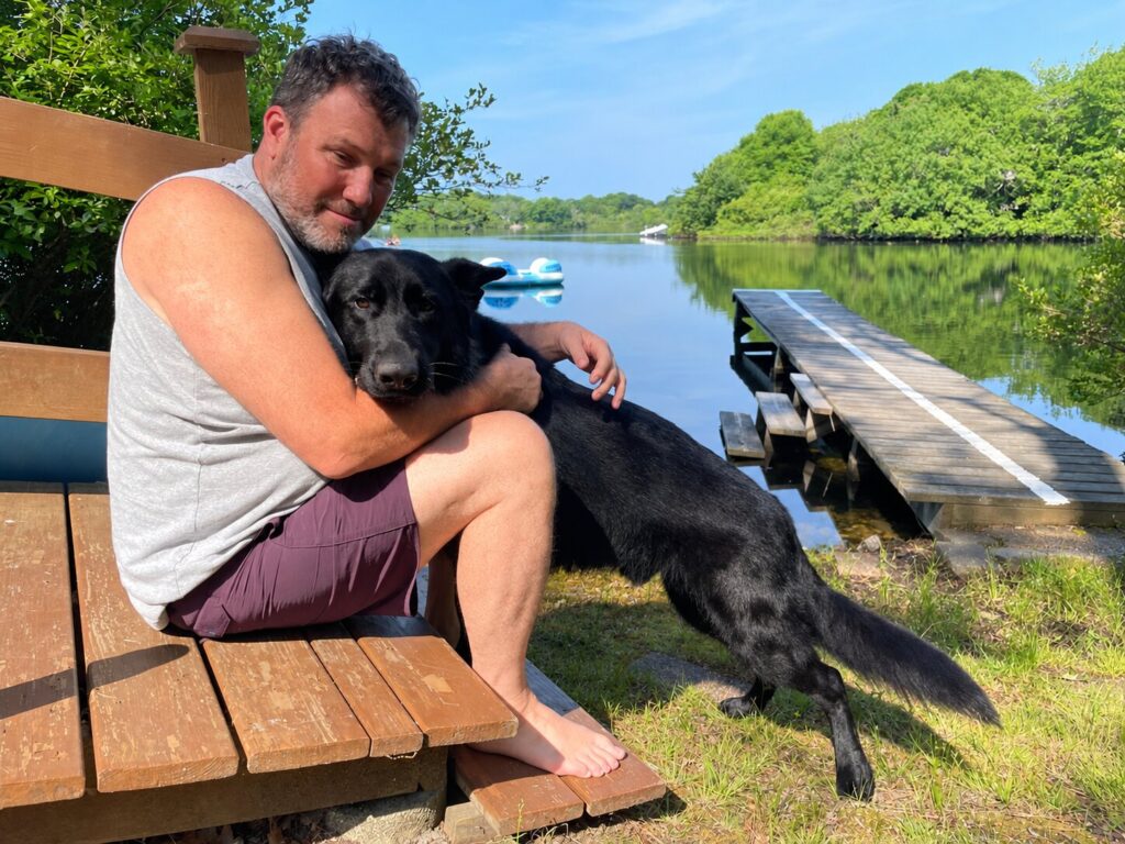 A man sitting on wooden steps hugs his black German Shepherd guide dog as the dog leans into him, with a calm lake and wooded shoreline in the background.