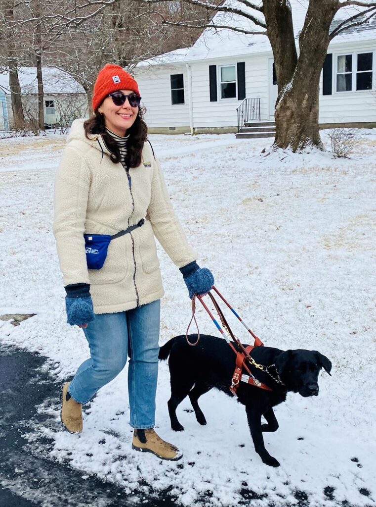 A smiling young woman wearing a cream coat and tan boots walks along a snowy country road with her black Lab guide dog.