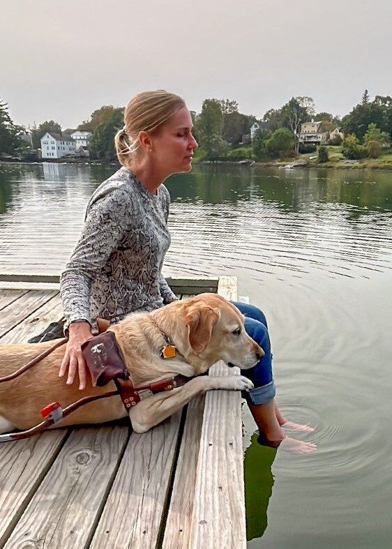A woman sits on a wooden dock with her feet in the water, resting an affectionate hand on her yellow Lab guide dog as they gaze across the water.