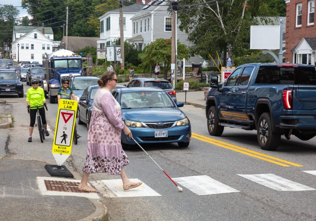 A woman using a long white cane crosses a busy street as a line of traffic and two cyclists wait.