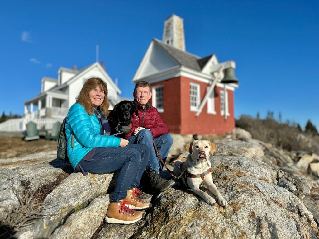 A couple sits on the rocks with their black and yellow Lab guide dogs near Pemaquid Point Lighthouse on the Maine coast.