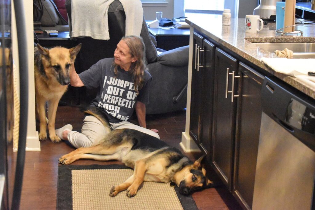 Sue sits on the kitchen floor, smiling as she pets one German Shepherd while another lies stretched out beside her on a rug.