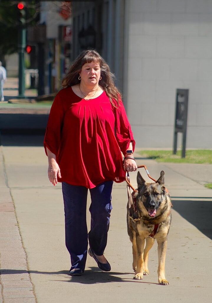 A woman wearing a bright red top walks along a city sidewalk with her German Shepherd guide dog.