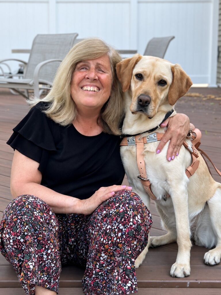 A woman smiling joyfully sits on a deck with her arm around a yellow Lab guide dog.