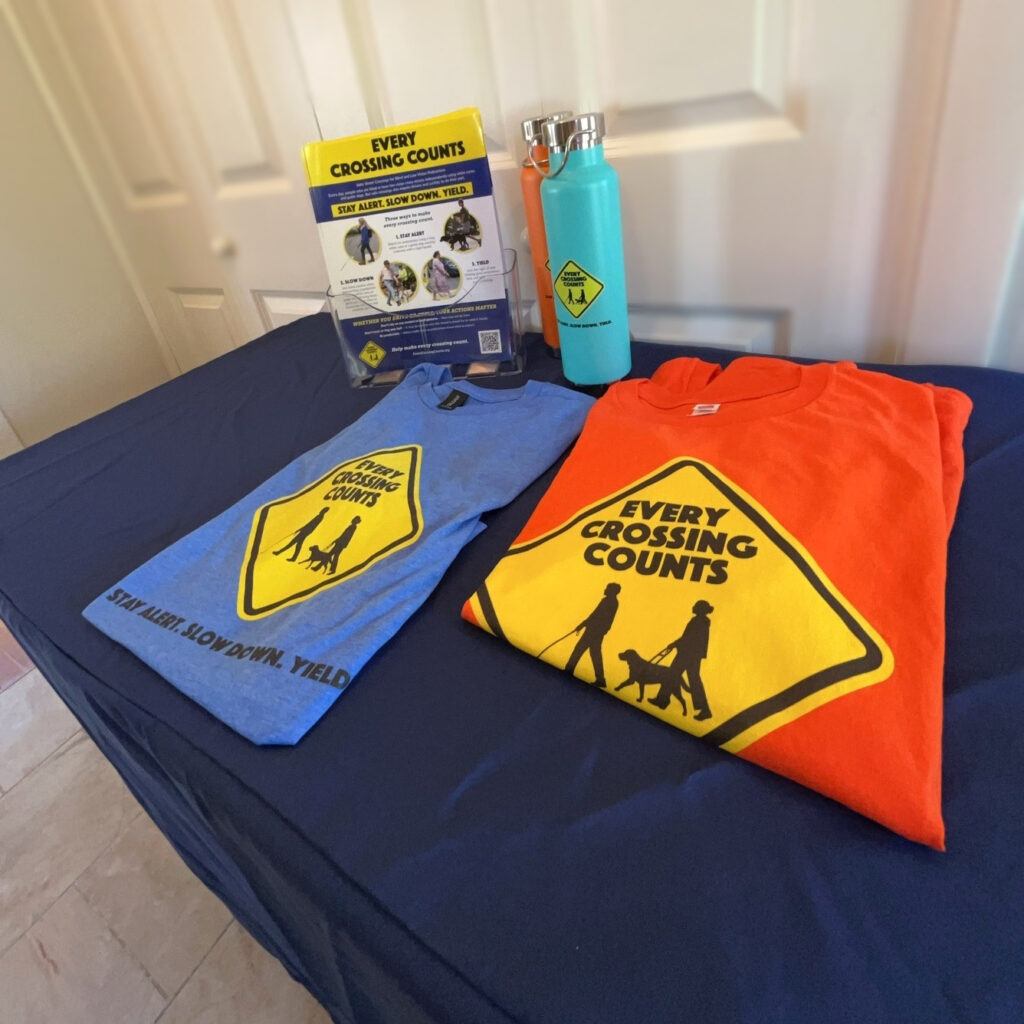 Every Crossing Counts display on a navy tablecloth, featuring blue and orange T-shirts, reusable bottles, and campaign flyers displayed in a clear, upright holder.